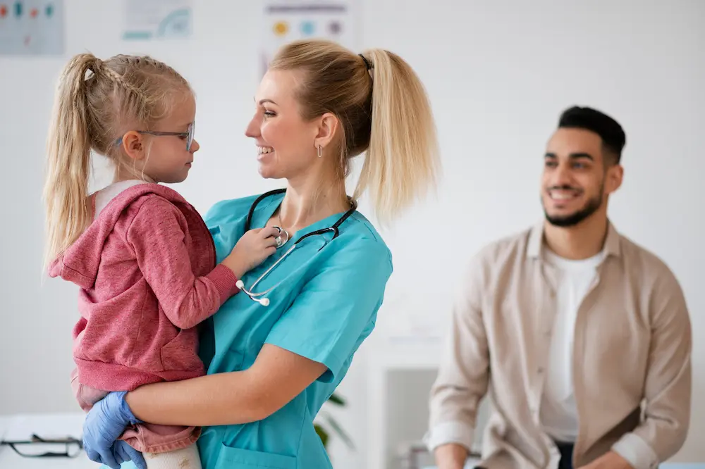 In a clinical setting, a smiling female doctor holds a young girl in the foreground. The child playfully interacts with the doctor’s stethoscope, while a man in the background looks on with a smile
