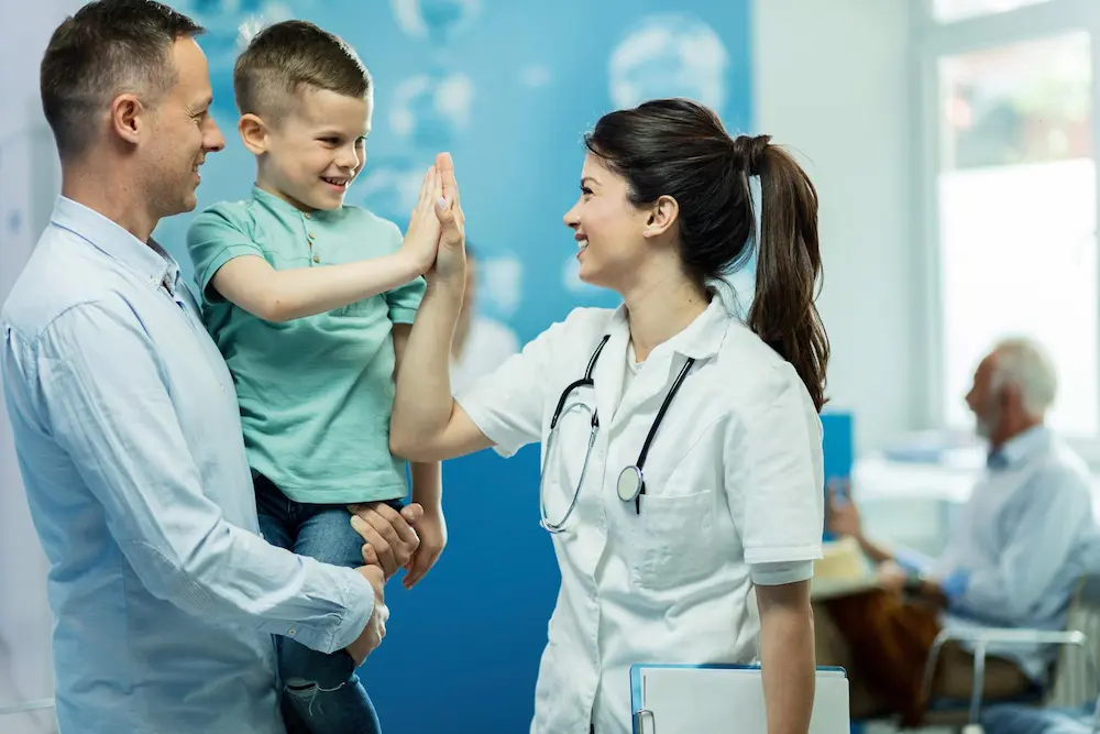 In a doctor's office waiting room, a father carries his young son, who is high-fiving a smiling female doctor. In the background, a blurred older man is seated, though his actions are indistinct and not central to the scene.