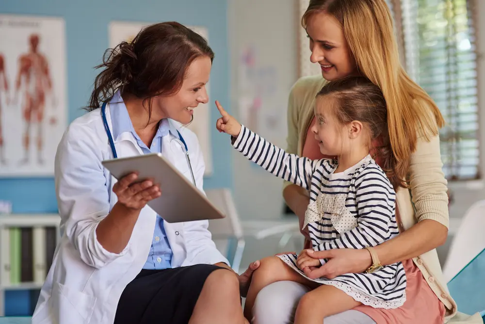 A doctor, mother, and daughter sit together in a clinical setting, all smiling. The daughter, held in her mother’s lap, reaches up with her right index finger toward the doctor’s nose, as if playfully engaging. The doctor holds up a clipboard, suggesting they had just been reviewing something.