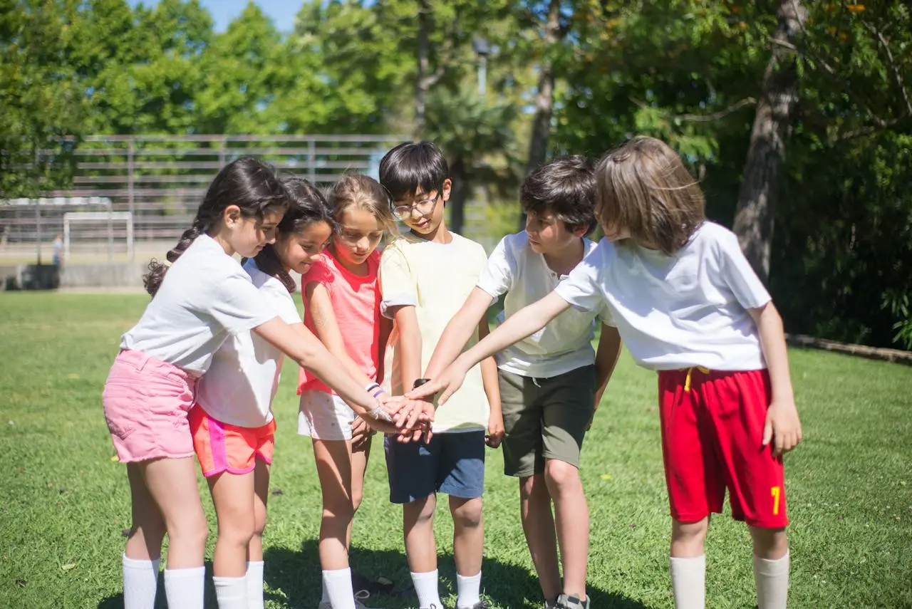 A group of children stand in a semi-circle on a grassy field, placing their hands together at waist height in a team huddle, symbolizing unity and teamwork.