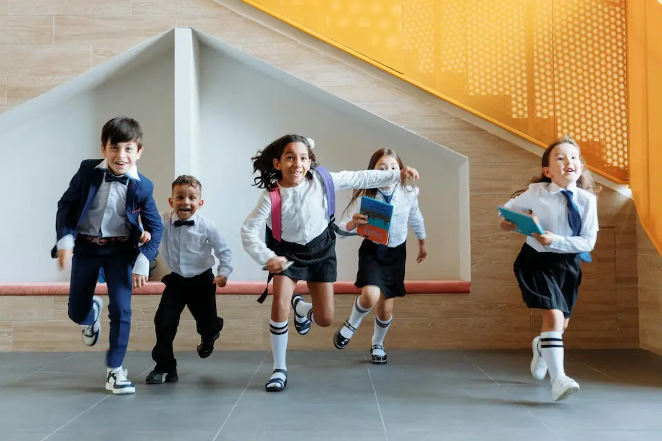 A group of children in school uniforms run excitedly down a hallway toward the camera, full of energy and enthusiasm.